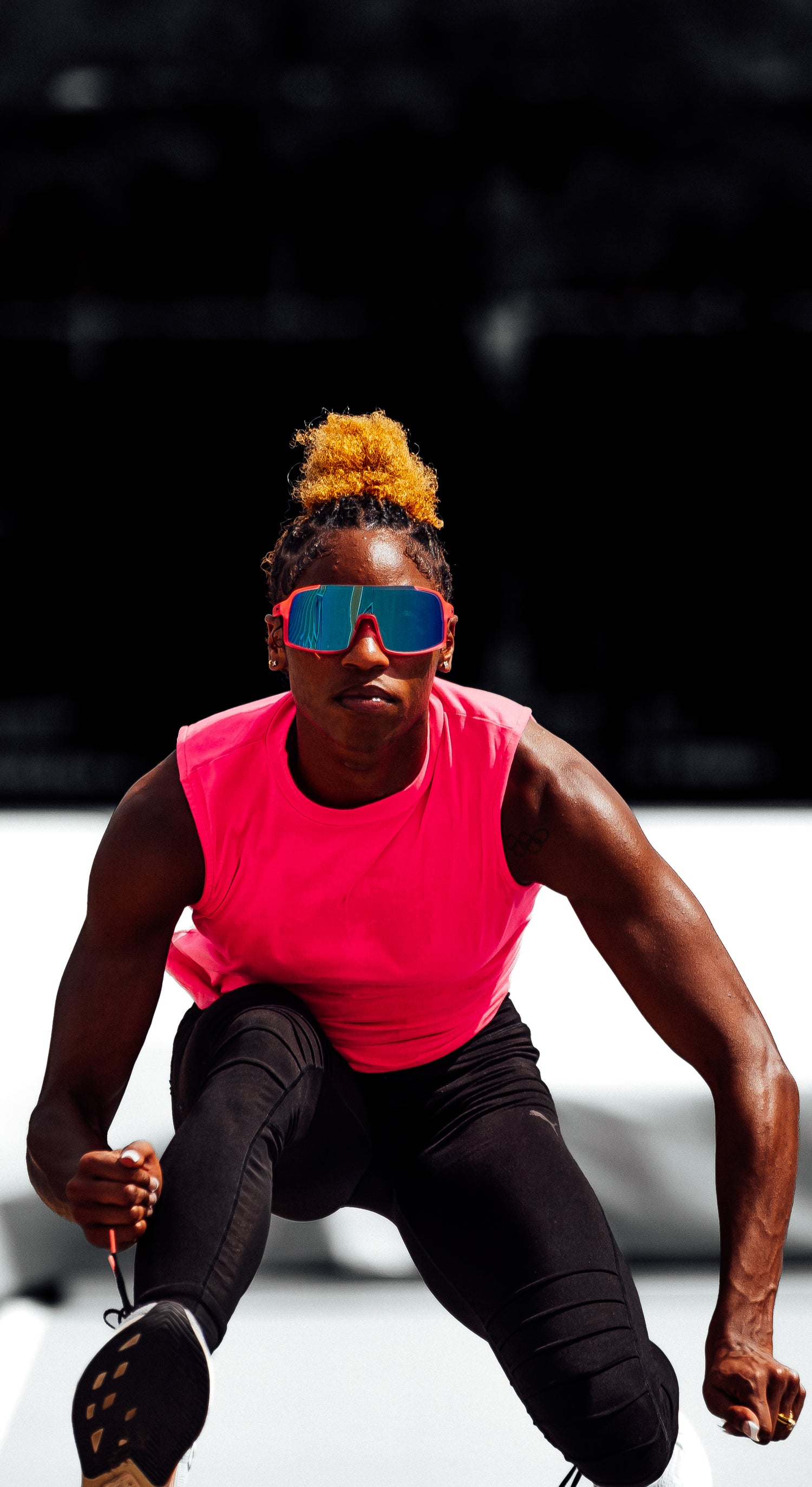 Person in a pink sleeveless top and black pants crouching with sunglasses on a blurred background