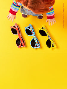 A doll with red hair and a colorful striped sweater is standing over three pairs of sunglasses arranged on a bright yellow surface. The sunglasses have different frame colors: red, blue, and yellow.