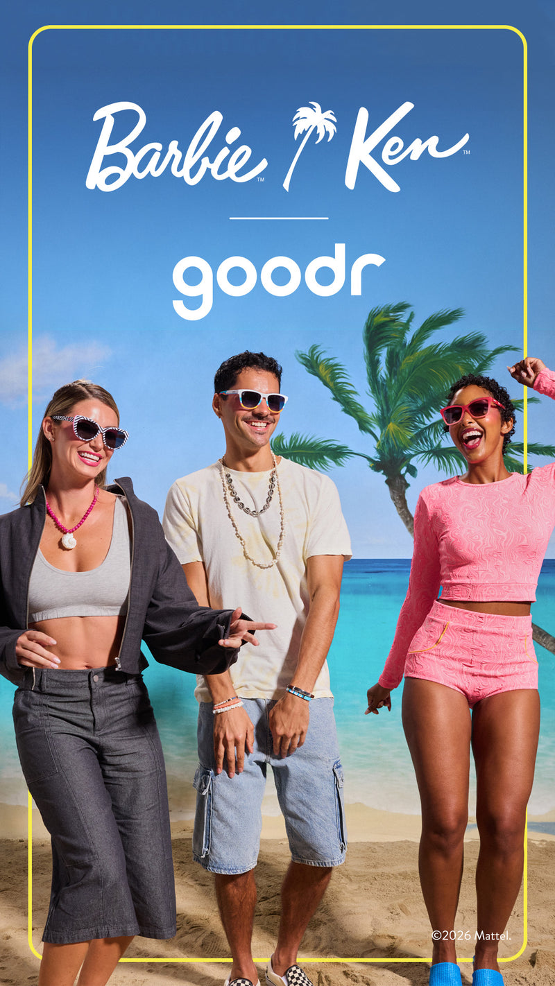 Three people walking on a beach with palm trees and ocean in the background, featuring the Barbie and Ken Goodr logo.