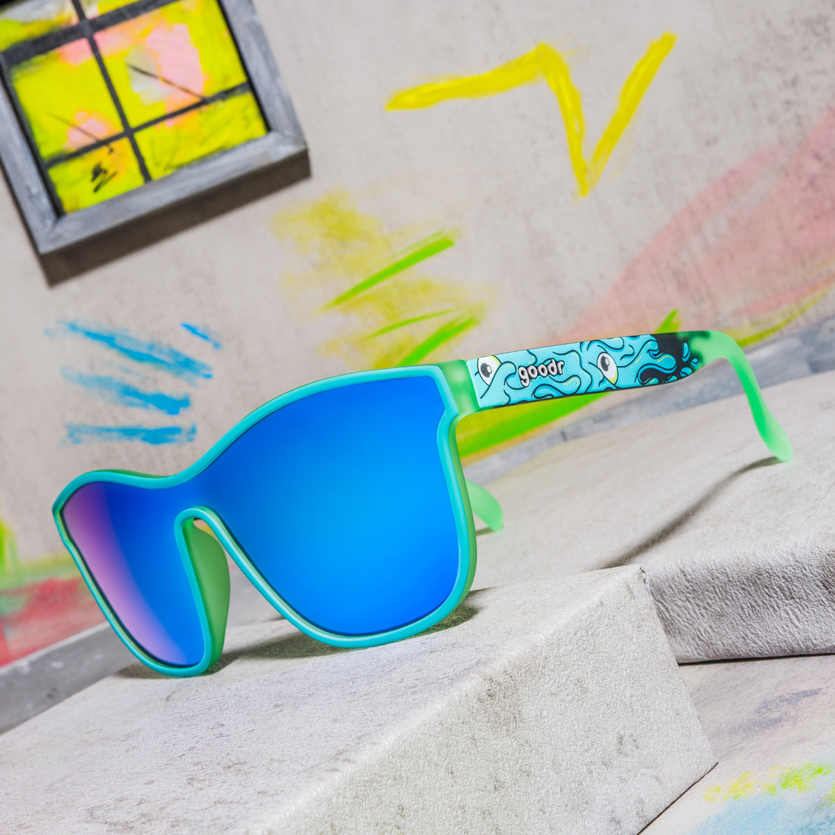 Mint green shield sunglasses with blue mirrored lenses and a wavy cartoon eye pattern on a colorful set.