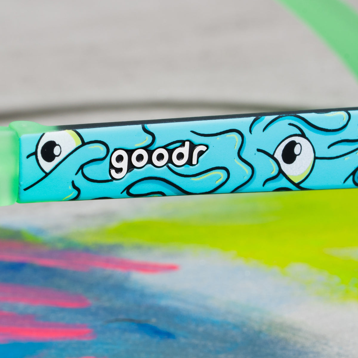 Close-up of the goodr logo on a mint green sunglass arm with a wavy cartoon eye pattern.