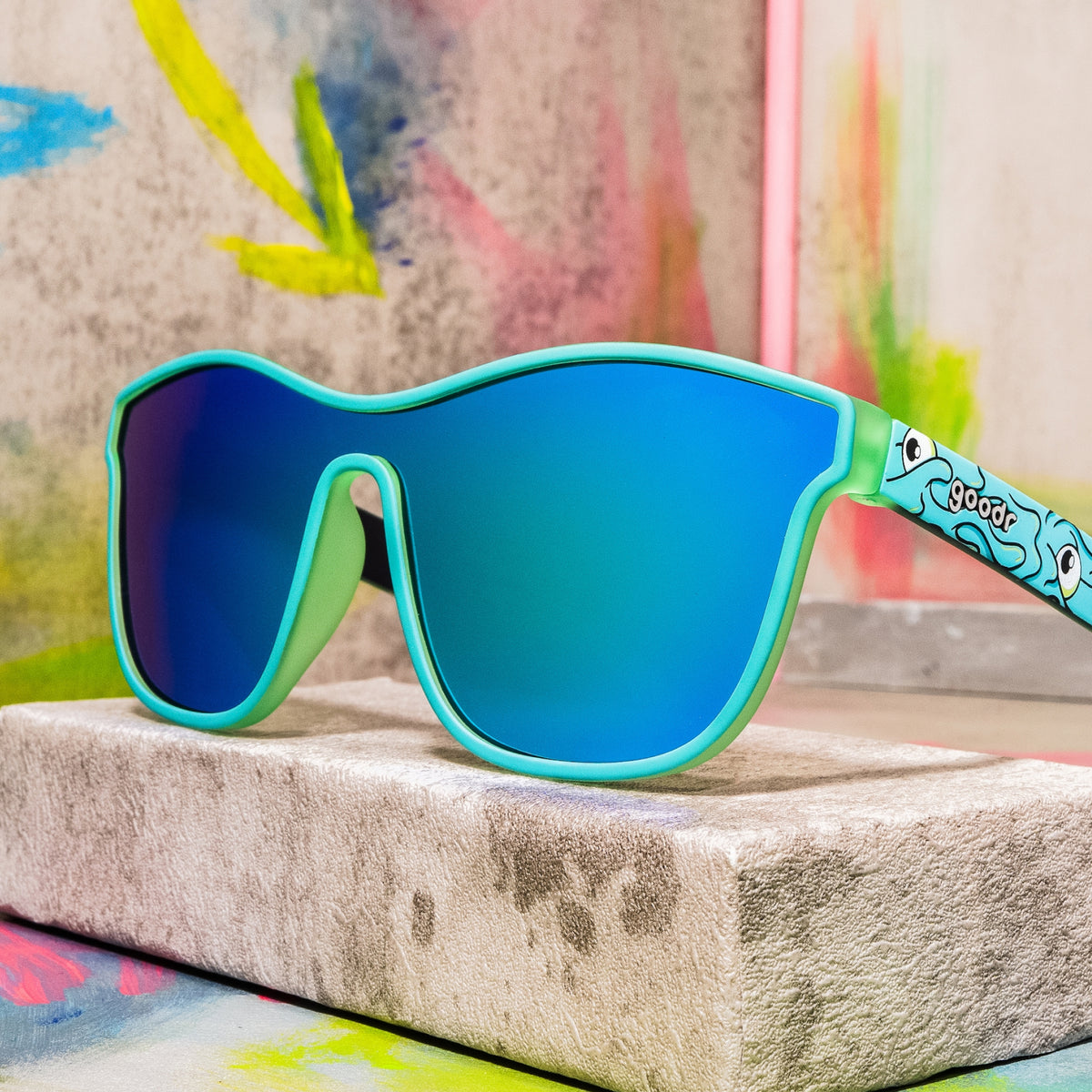 Mint green shield sunglasses with blue mirrored lenses and a wavy cartoon eye pattern on a concrete block.