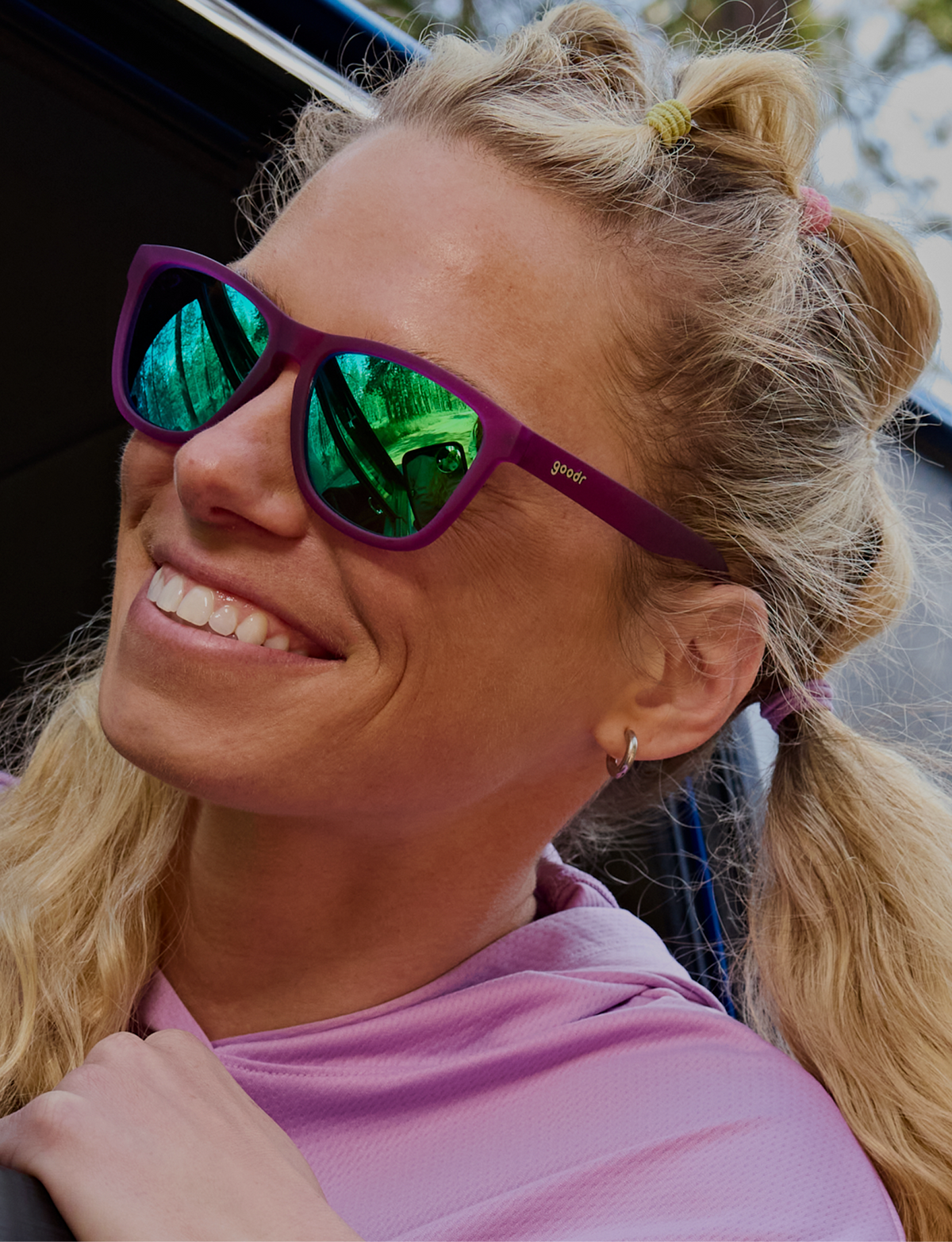 A close-up of a woman wearing purple sunglasses with green mirrored lenses and a matching purple shirt.