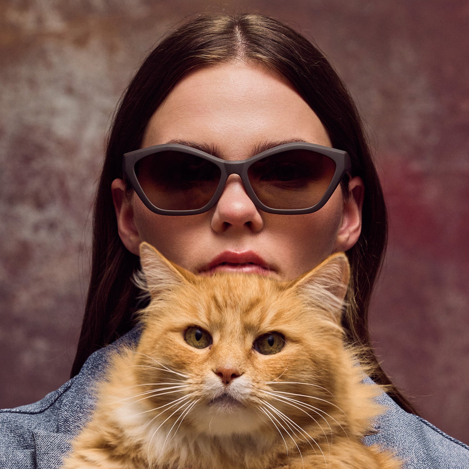 Woman wearing sunglasses with a cat on her shoulder against a brown background