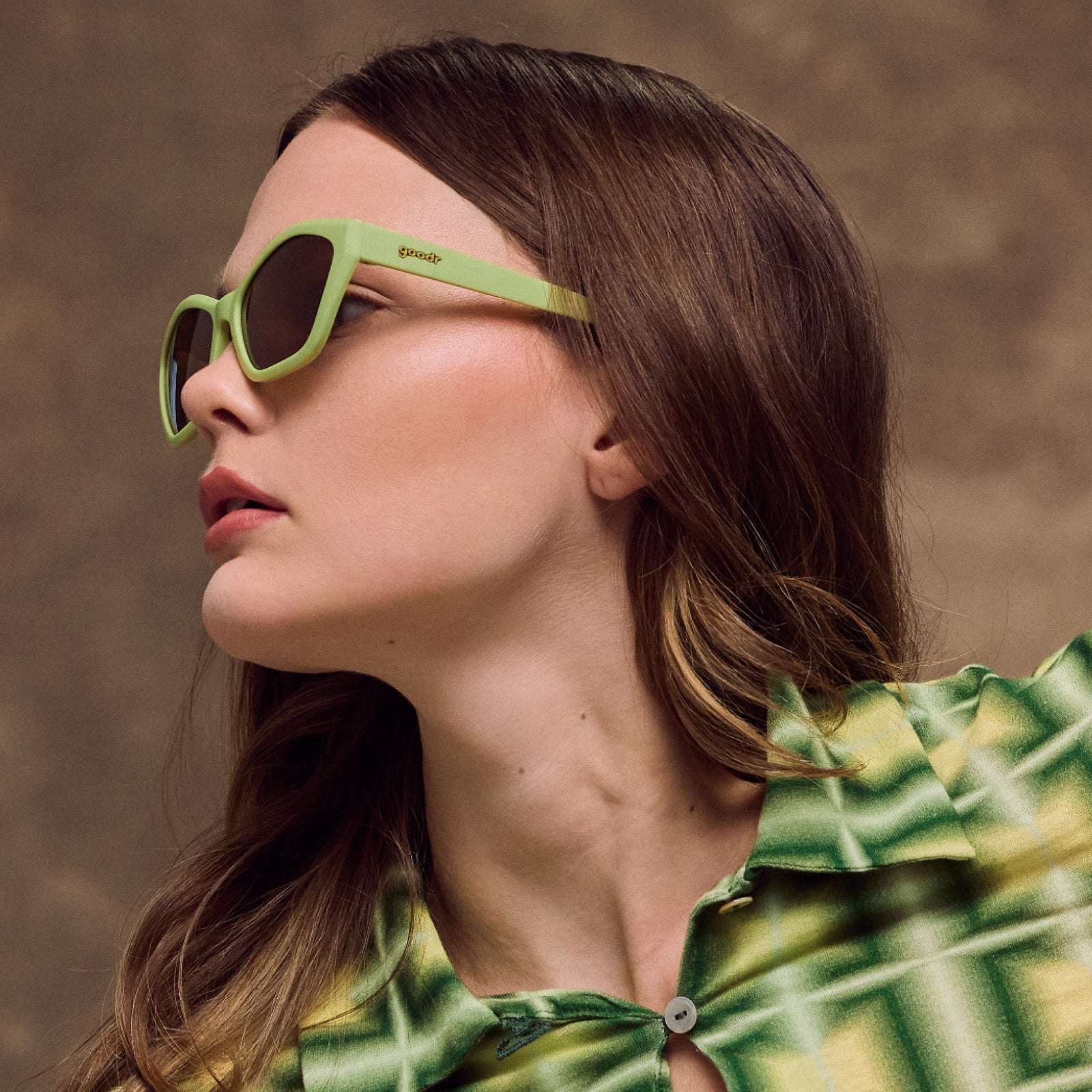 A woman poses wearing green cat-eye sunglasses featuring a geometric frame and brown lenses.