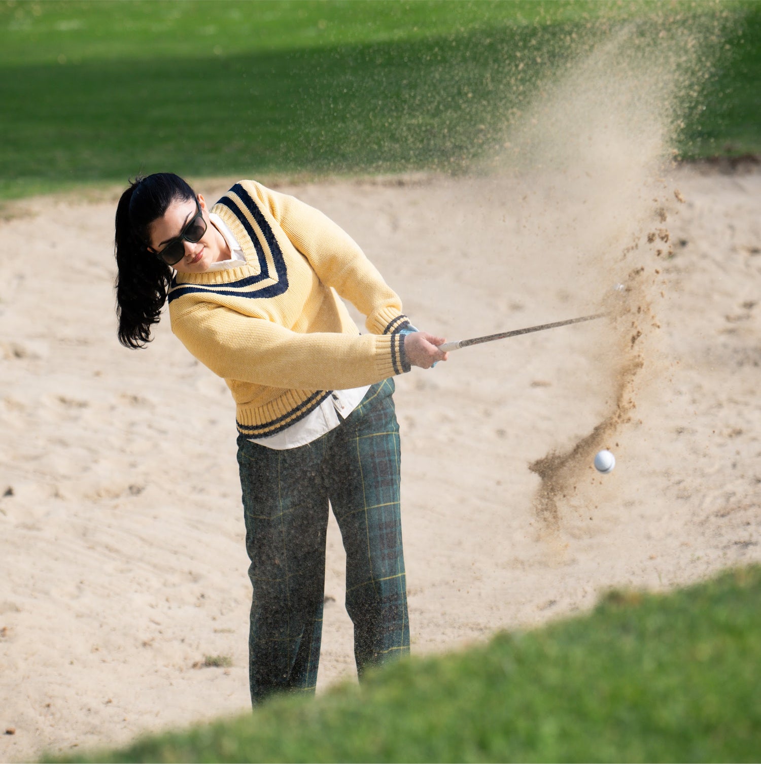 Person playing golf, hitting a ball out of a sand trap on a green course.