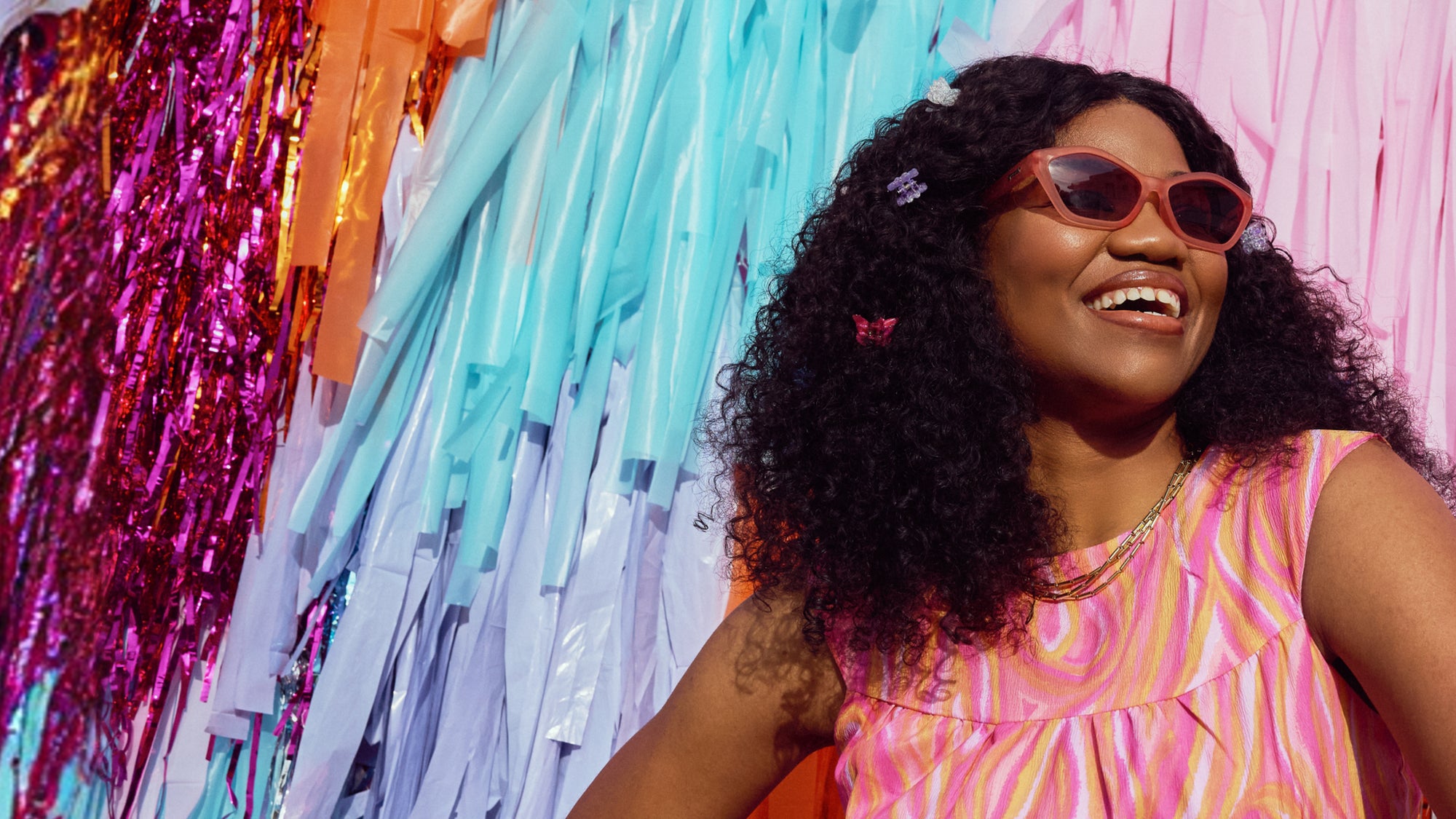 Woman wearing sunglasses and a colorful dress in front of a vibrant, multicolored curtain.