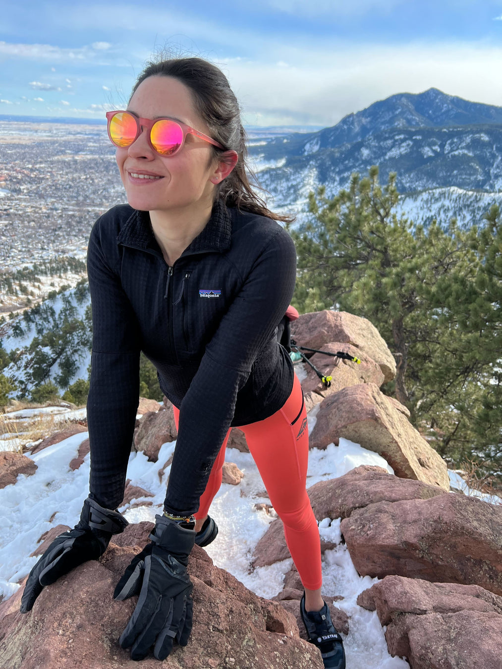 Woman in athletic clothing standing at the top of a mountain, wearing round, pink sunglasses with sunset-colored lenses