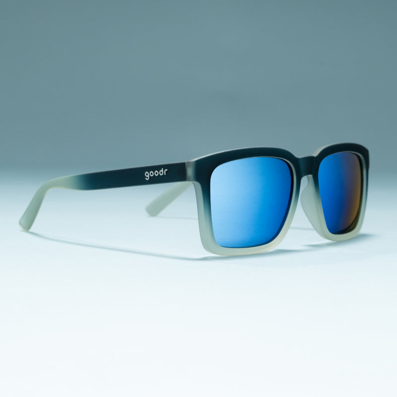 Angled view of translucent black & teal shades w/ blue reflective lenses+ a classic square silhouette on a flat surface.
