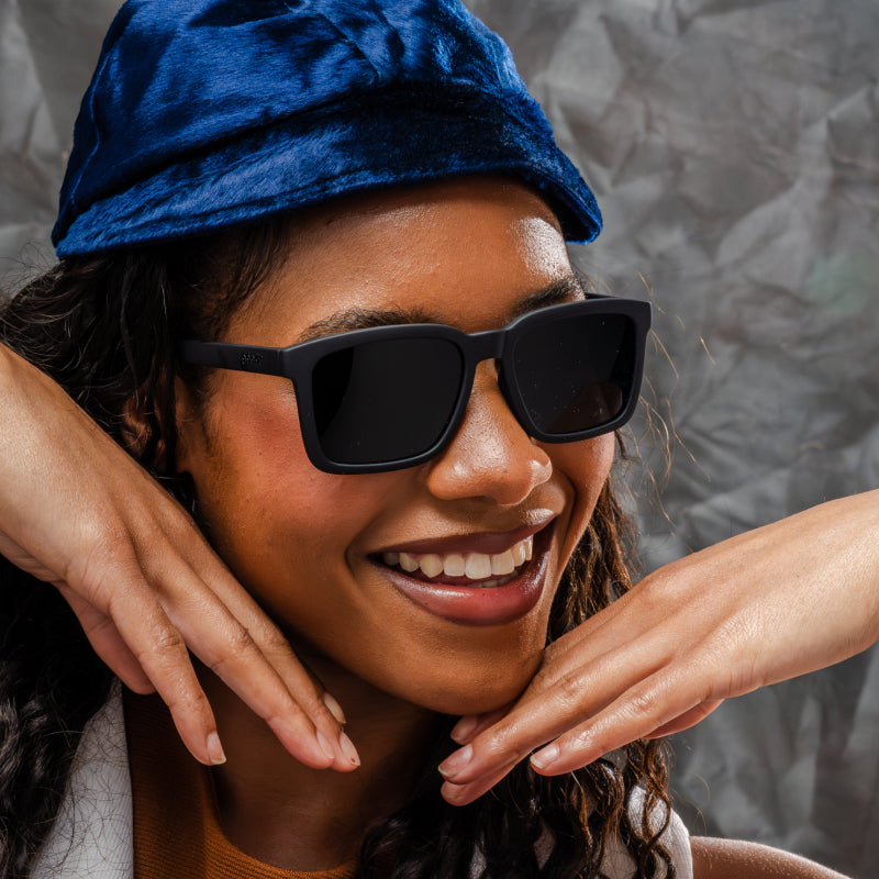 A woman is smiling & posing in black sunglasses with a classic square silhouette & black nonreflective lenses.