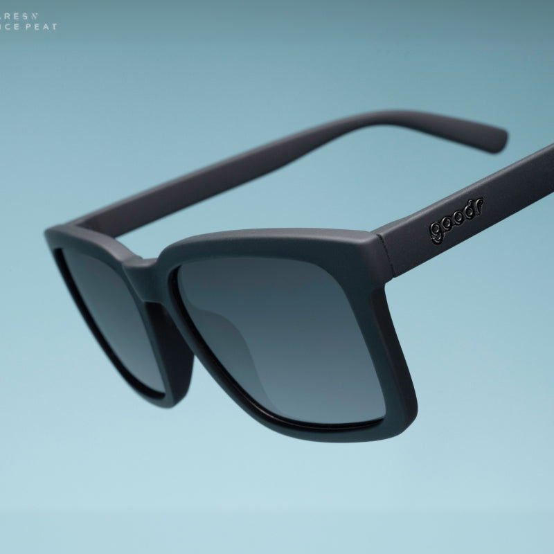 Three-quarter angle close-up of black sunglasses with a square silhouette & black nonreflective lenses tilted down.