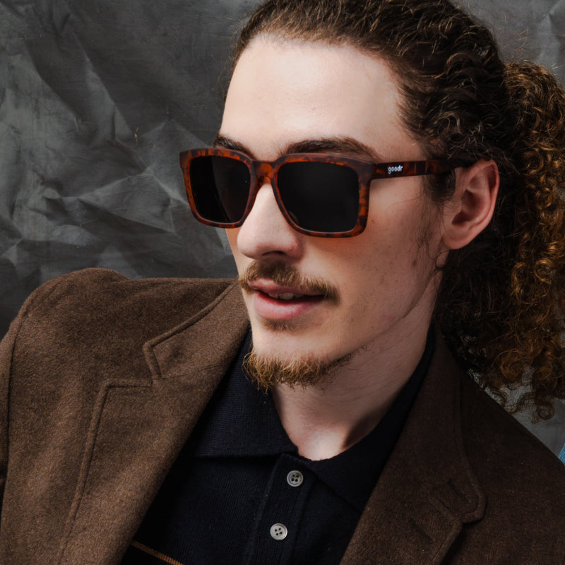 A man with a ponytail wearing translucent brown square shades w/ a tortoiseshell pattern & brown nonreflective lenses.