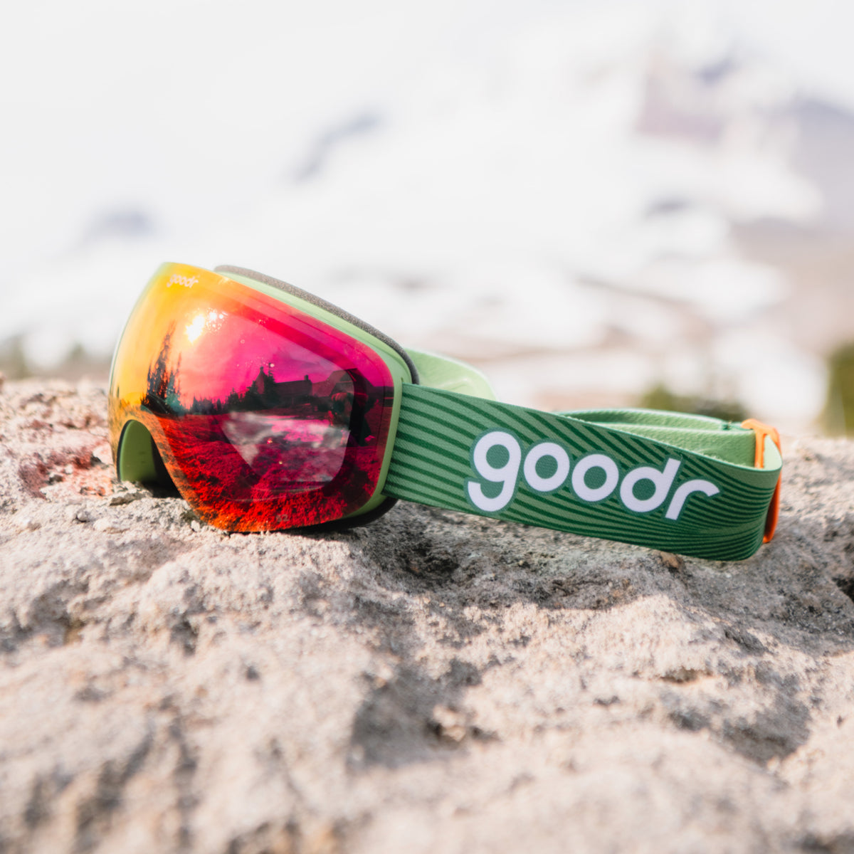 Smoke red mirrored snow goggles with a green frame and green goodr strap resting on a rock.