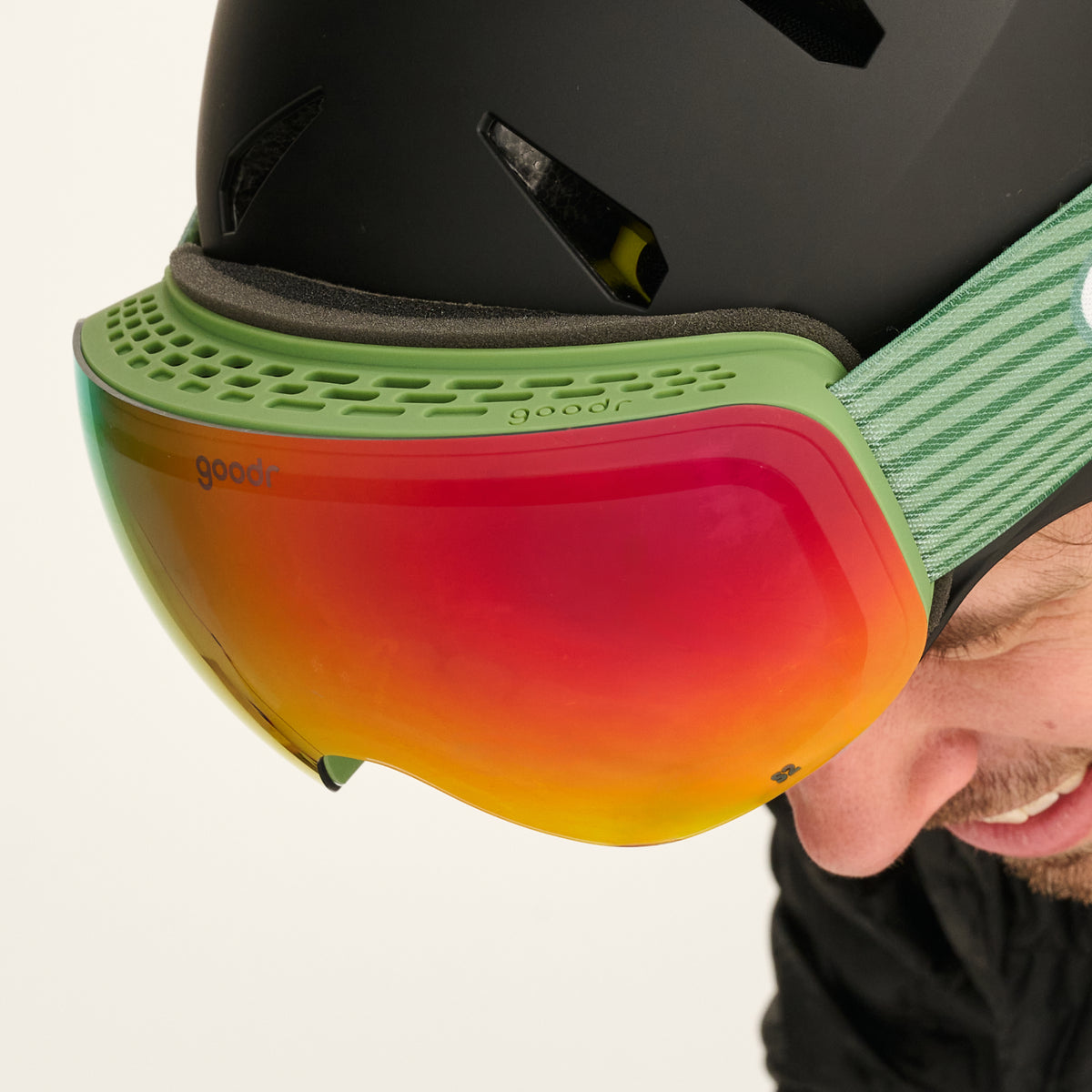 Close-up of smoke red mirrored snow goggles with a green vented frame worn with a black helmet.