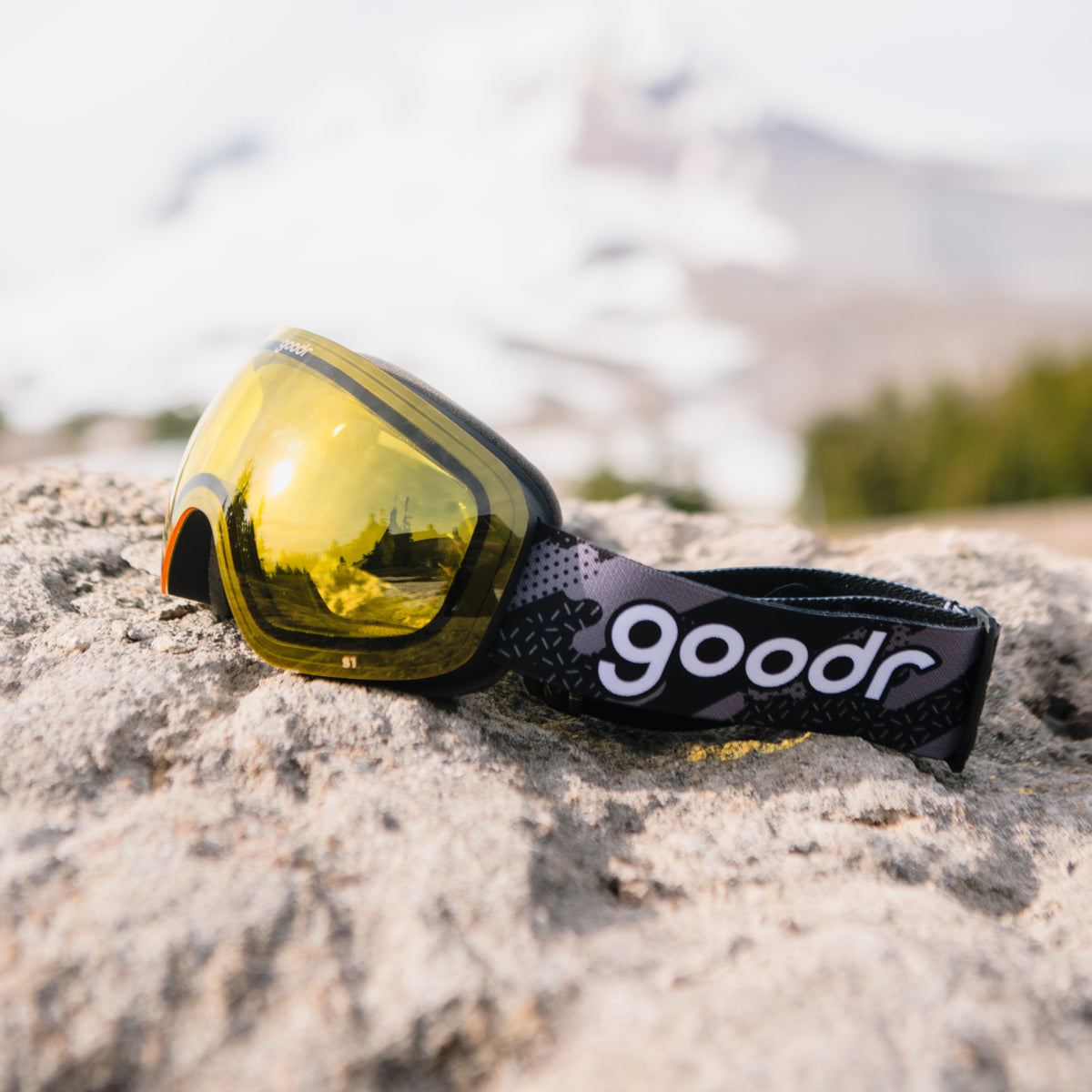 Yellow snow goggles with a black frame and patterned goodr strap resting on a rock.
