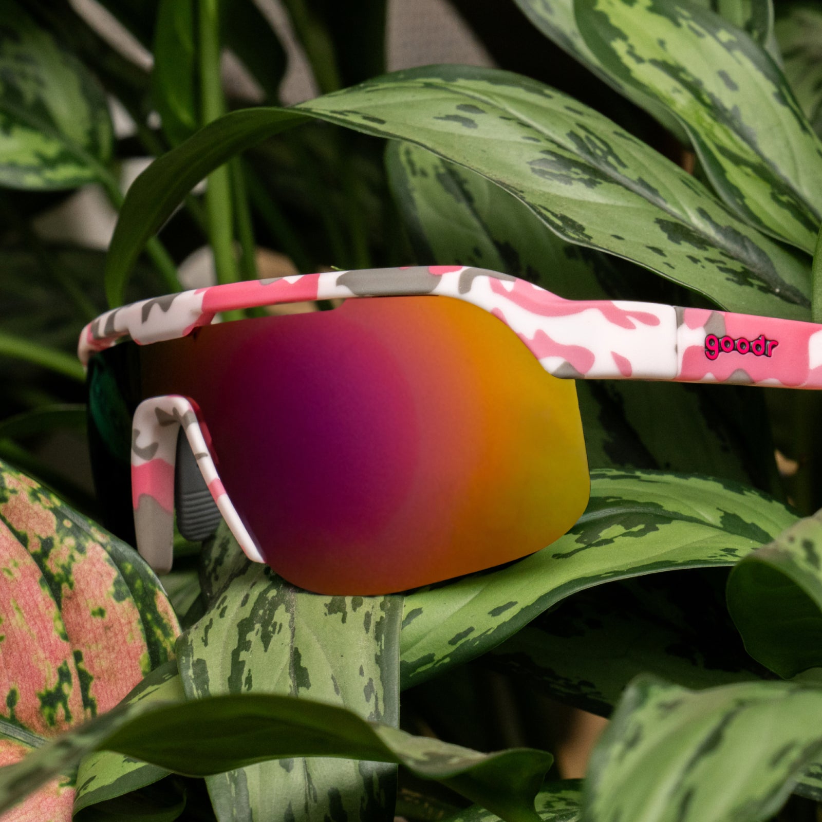 Pink and white camouflage wrap-around sunglasses with a mirrored pink, orange, and yellow gradient lens, resting on green tropical leaves. The brand name 'goodr' is visible on the temple arm.