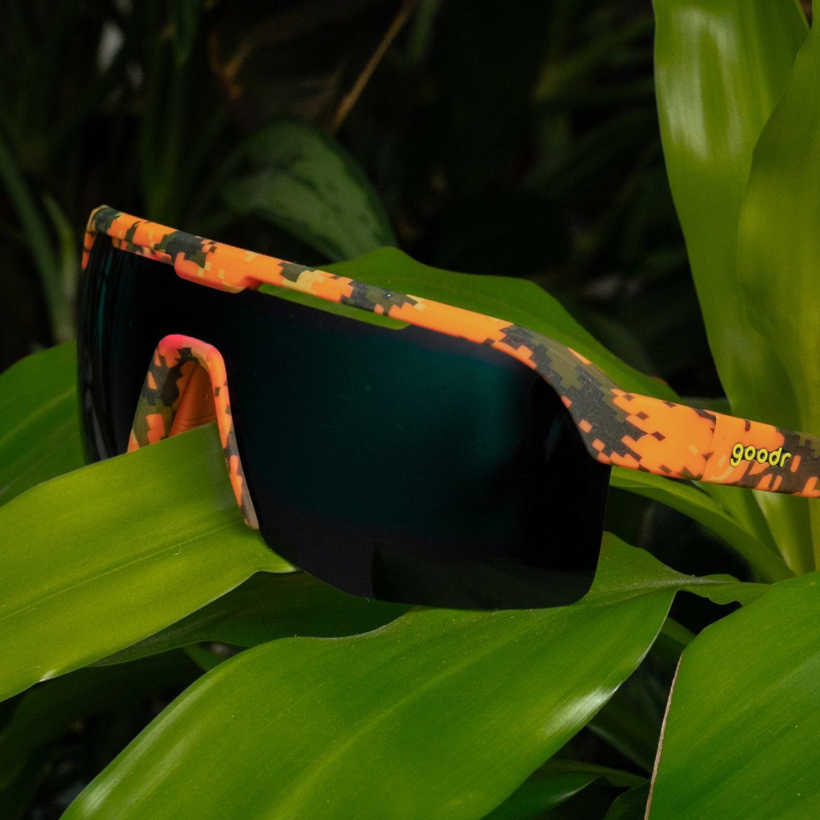 Orange camouflage sport sunglasses with dark gradient lenses resting on bright green tropical leaves. The brand name 'goodr' is printed in yellow on the temple arm.