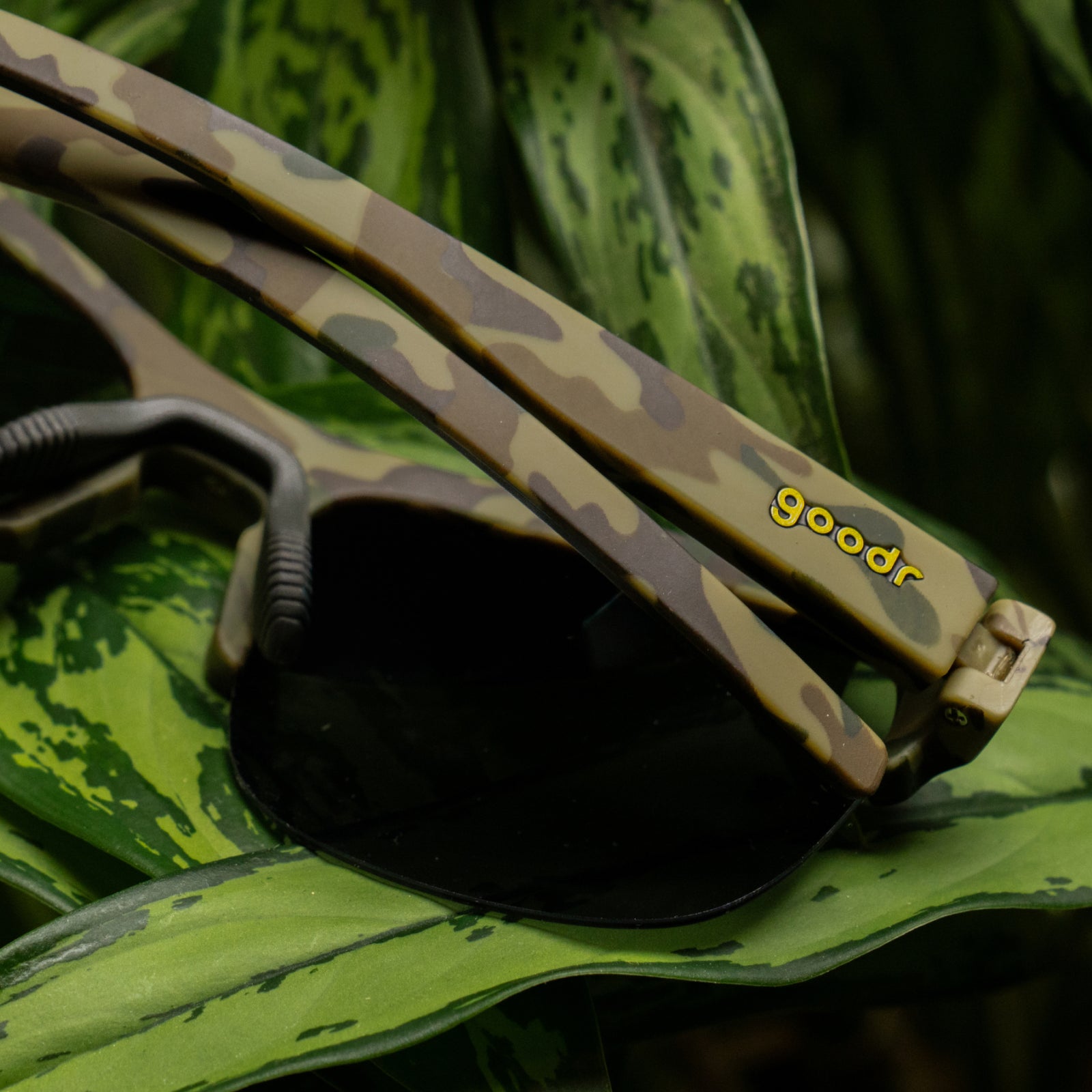 Close-up of camouflage-patterned sunglasses resting on green leaves. The frames are tan and olive green with a yellow 'goodr' logo, and the dark lenses are visible.