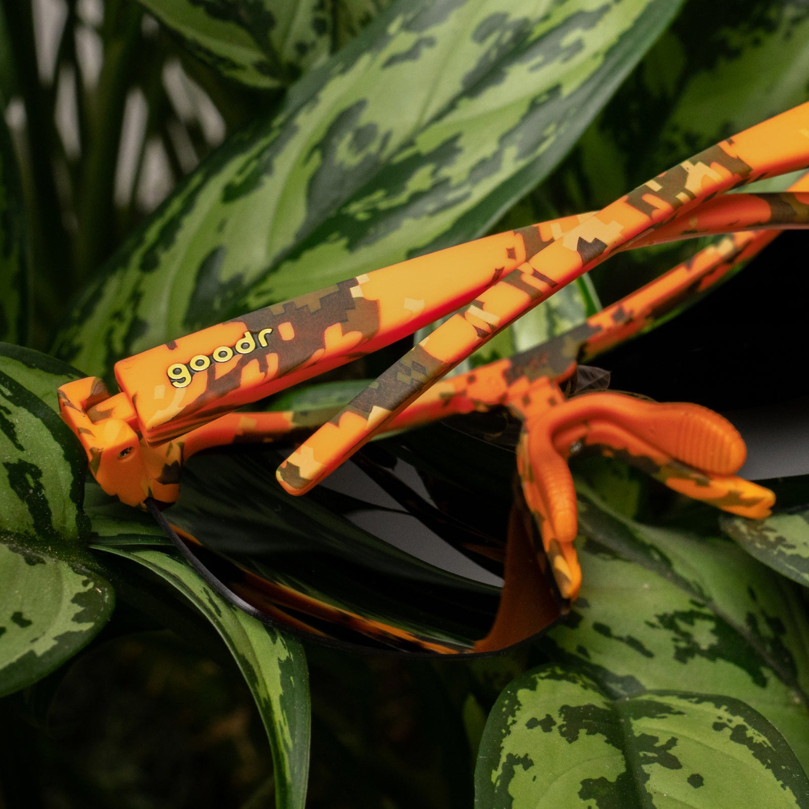 Orange and brown camouflage sunglasses with black lenses resting on green patterned leaves. The brand name 'goodr' is printed in yellow on the temple arm.