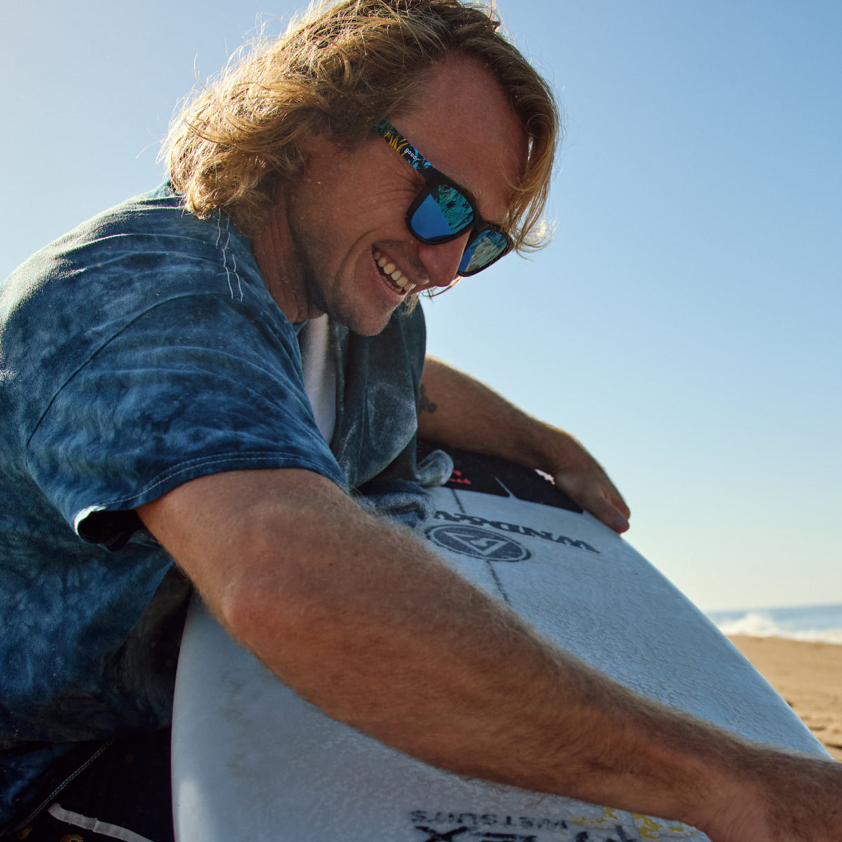 Ben Gravy wearing goodr sunglasses, holding his surfboard while smiling at the beach.