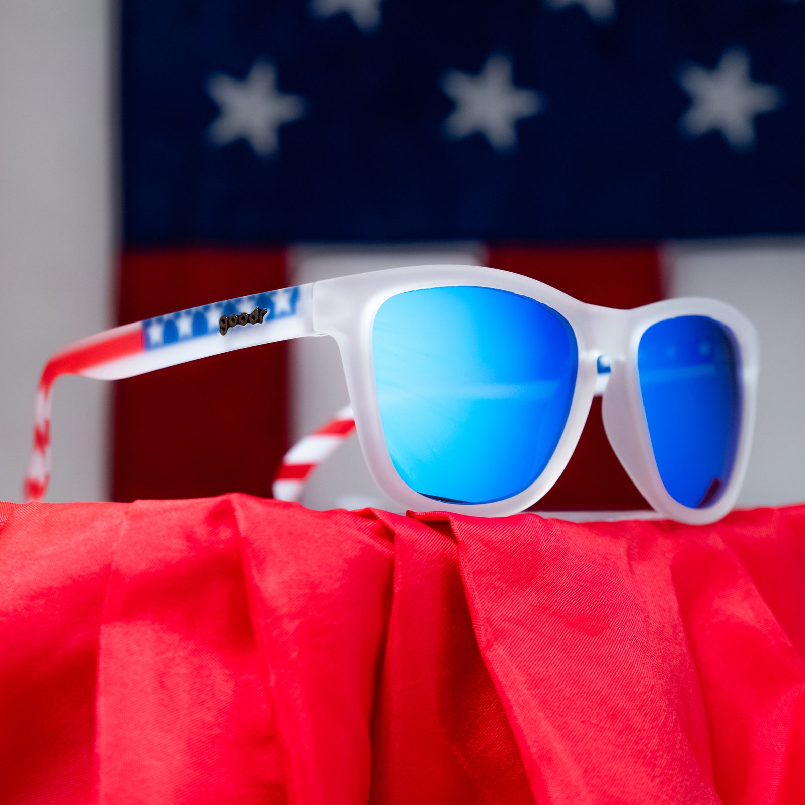 Angled view of clear shades w/ an American flag pattern+blue reflective lenses on a red podium w/ flag in background.