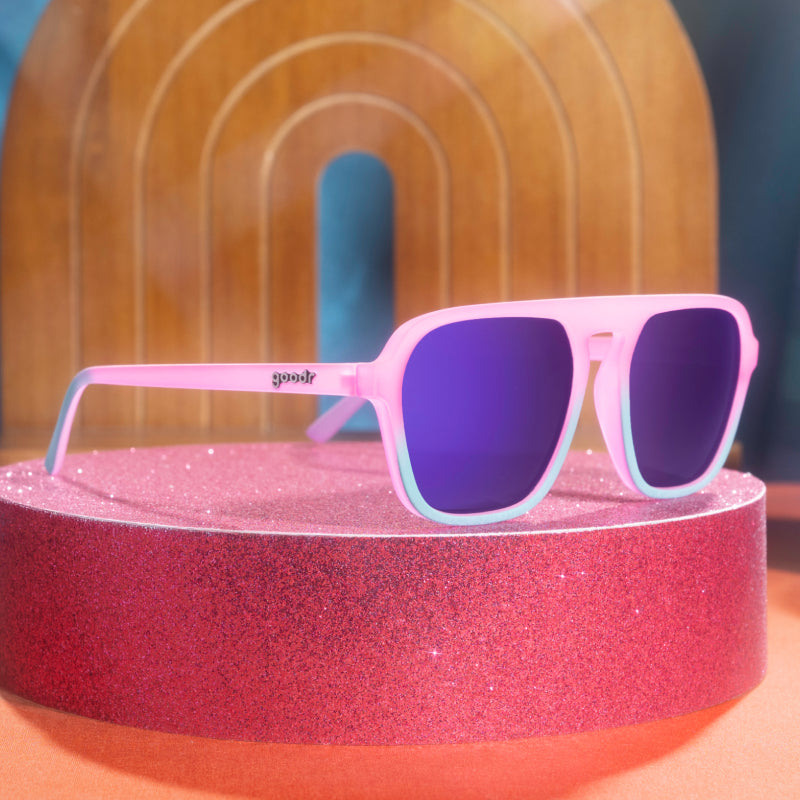 Pink and teal aviator sunglasses with reflective lenses on a pink glittery tabletop.