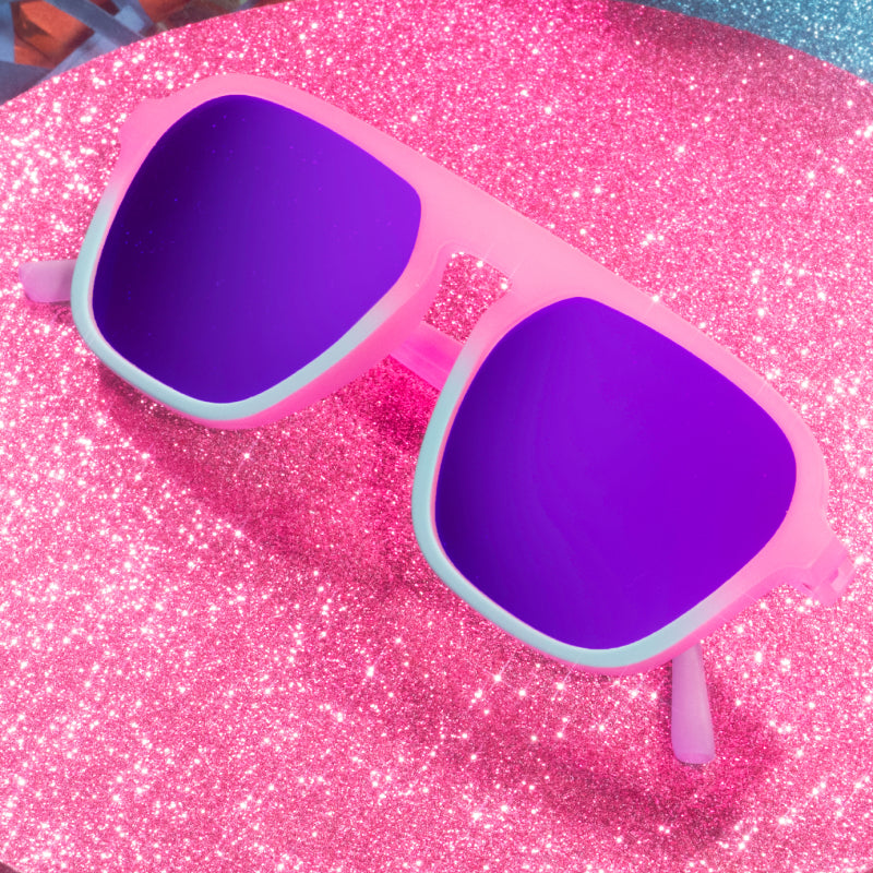 Pink and teal aviator sunglasses with reflective lenses folded on top of a pink glittery tabletop.