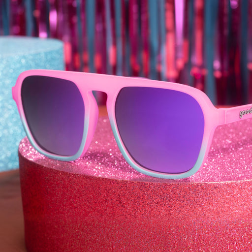 Pink and teal aviator sunglasses with reflective lenses on a glittery tabletop in front of a fringe glitter curtain in the back.