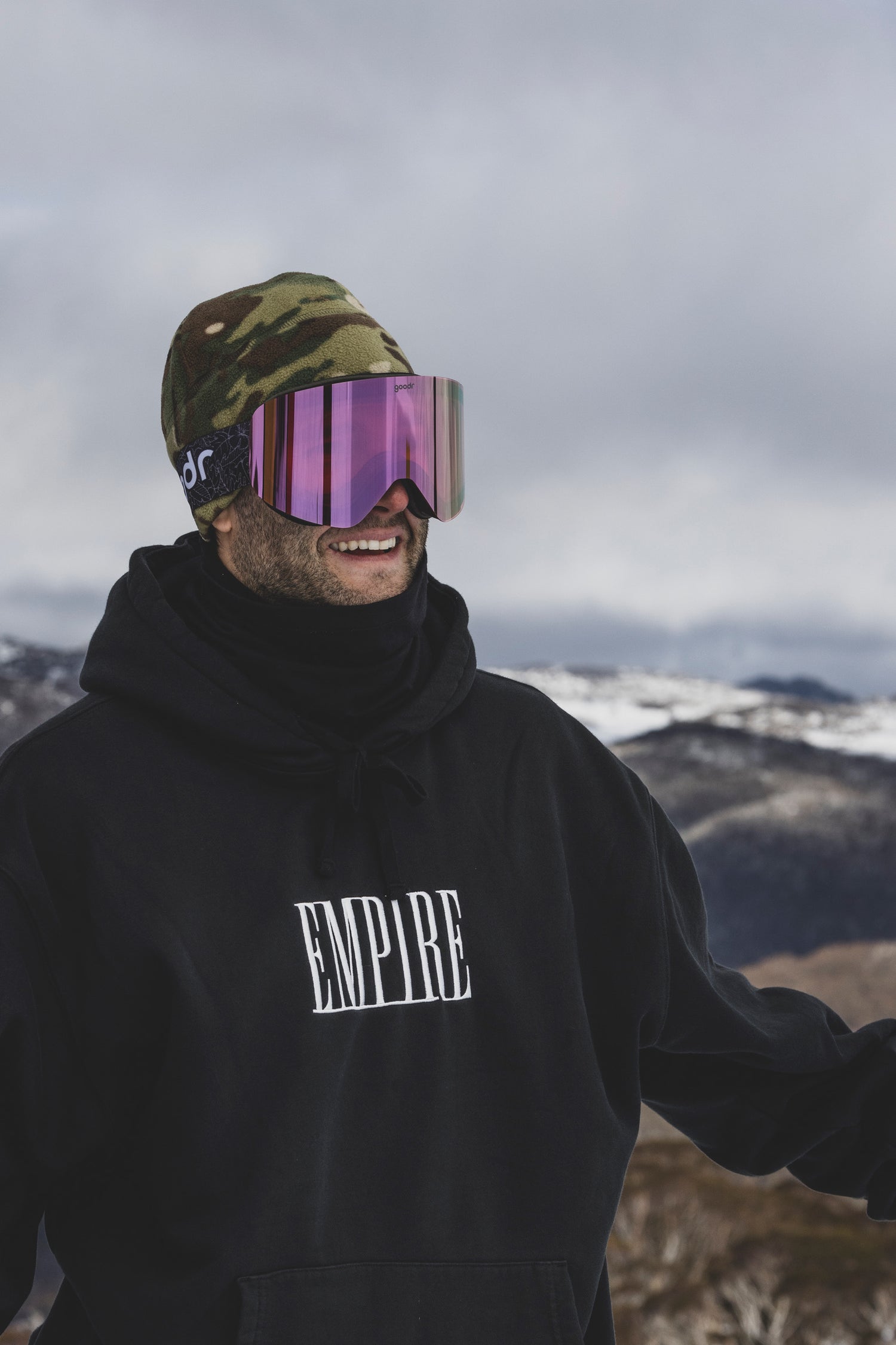 Person wearing a black hoodie, camouflage beanie, and purple goodr goggles in a mountainous landscape.