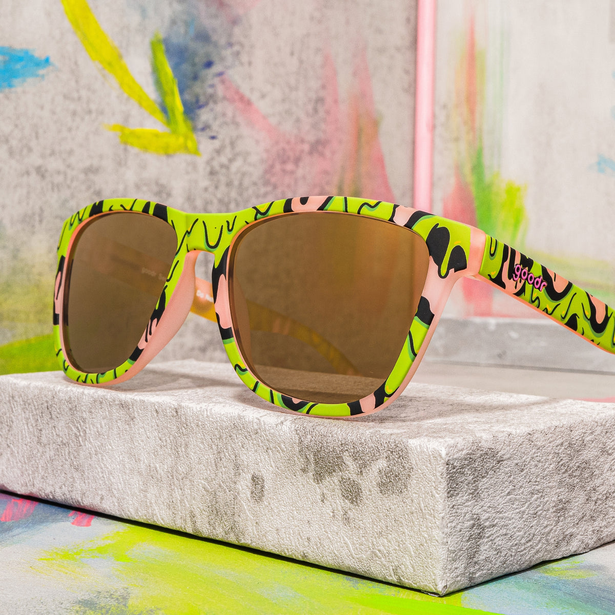 Pink sunglasses with gold lenses and a gooey green-and-black slime pattern resting on a concrete block.