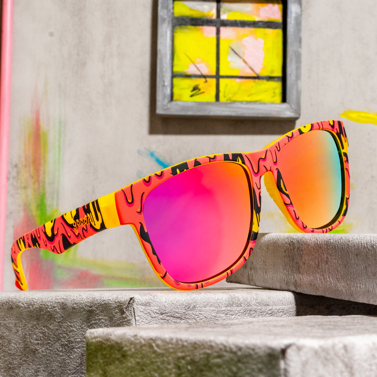 Goopy-patterned sunglasses with pink mirrored lenses resting on concrete blocks against a colorful painted backdrop.