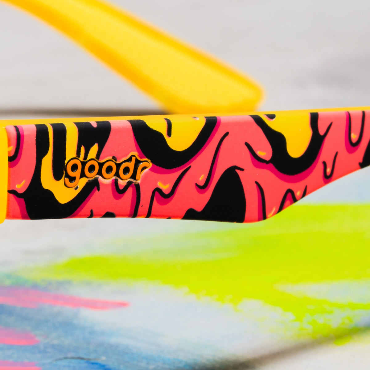 Close-up of the goodr logo on the arm of red, yellow and black goopy-patterned sunglasses against a colorful backdrop.