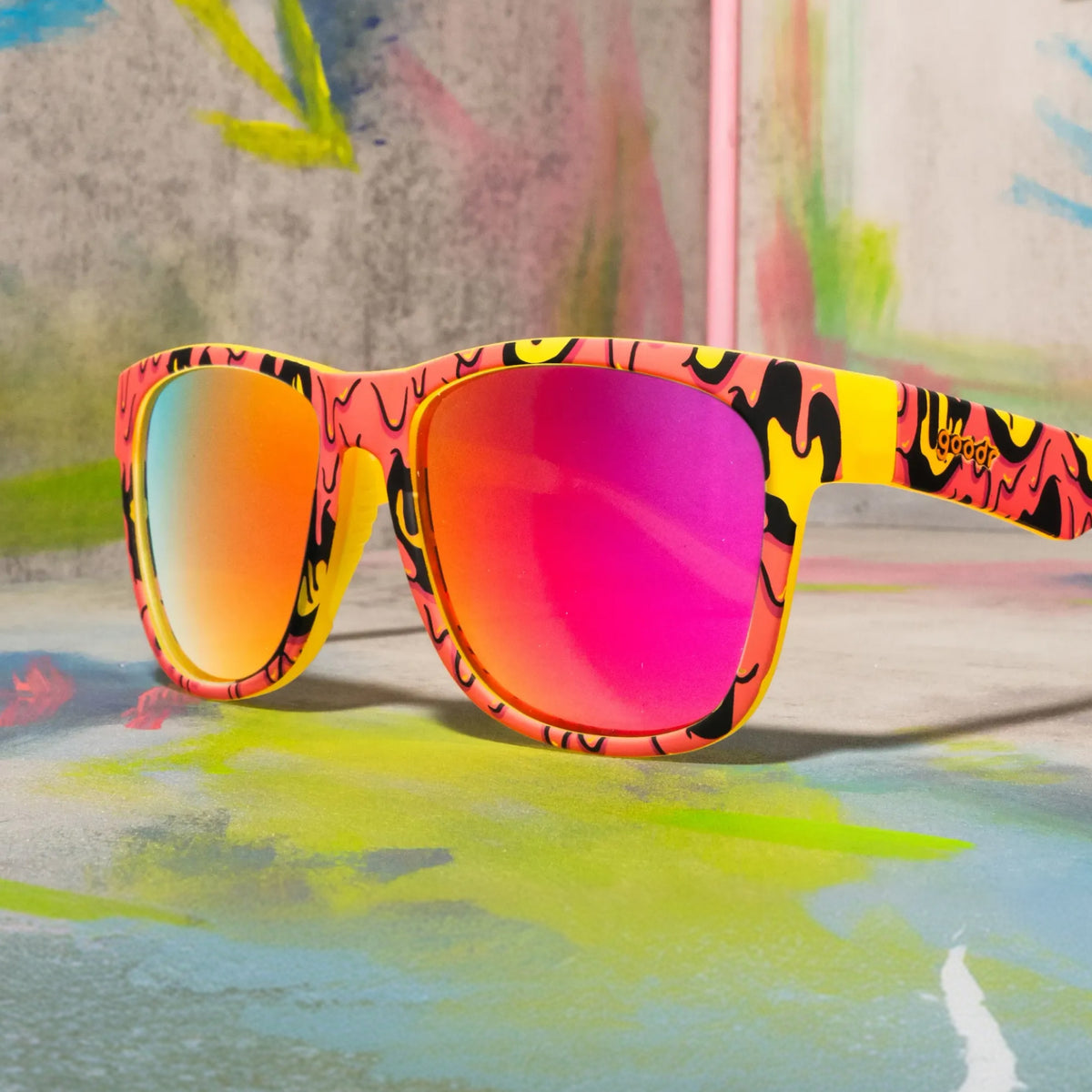 Goopy-patterned sunglasses with pink mirrored lenses resting on a colorful painted surface and backdrop.