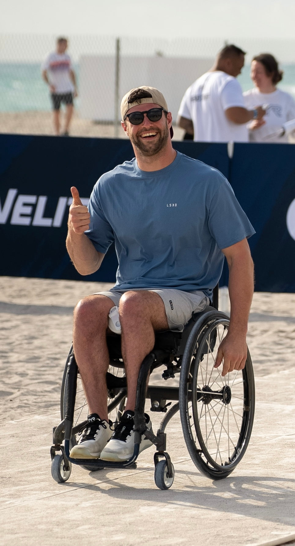 Man in a wheelchair giving a thumbs-up on a beach with people in the background.