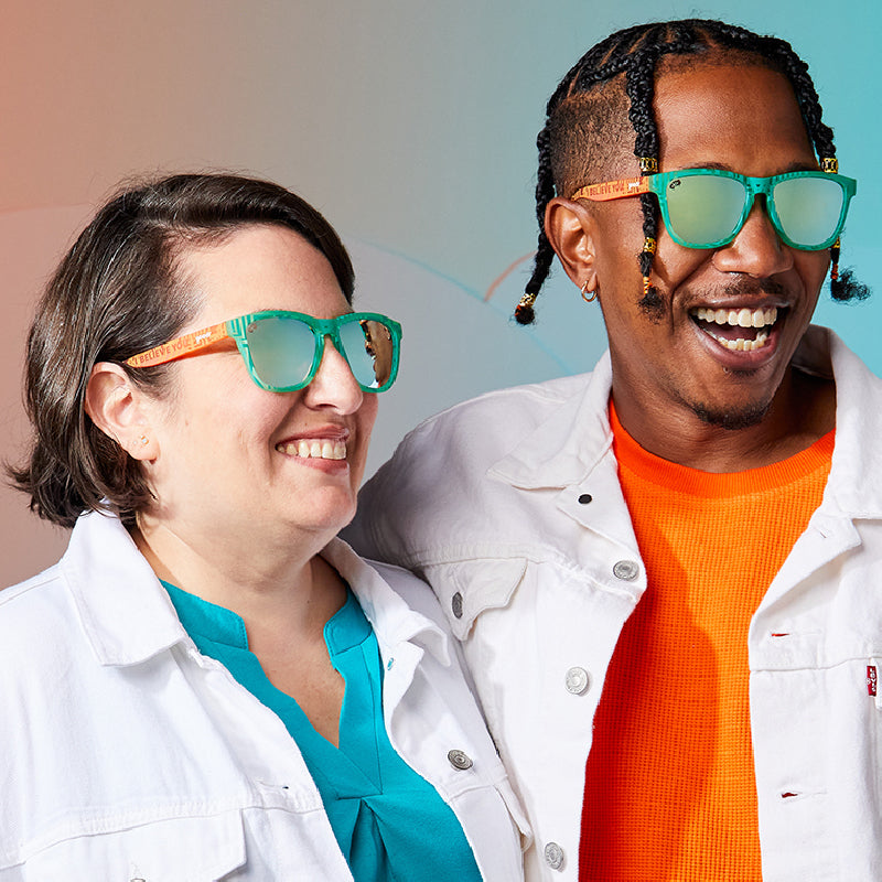 Two people side by side wearing sunglasses, one in a white jacket and turquoise top, the other in a white jacket over an orange shirt.