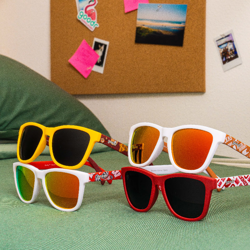 Four pairs of sunglasses are arranged on a bed with a green comforter. The sunglasses have yellow, red, and white frames, with different lens colors. A corkboard with photos and pink notes is visible in the background.