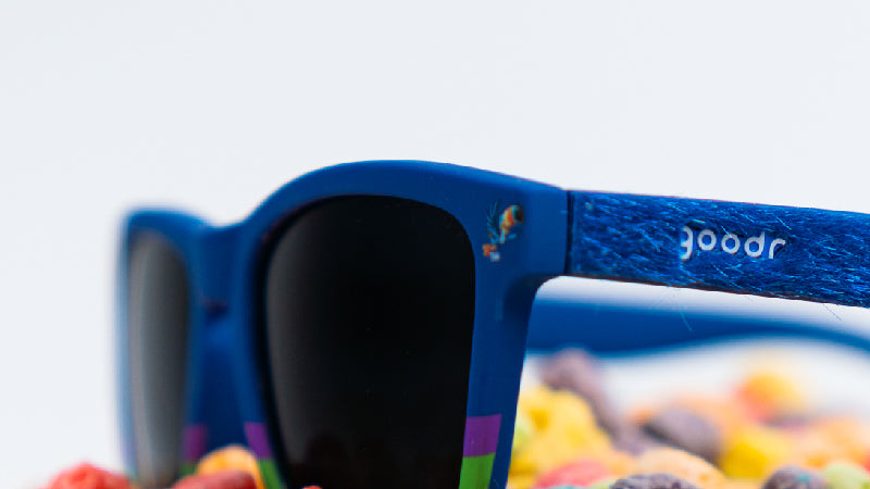 A detailed close-up of blue sunglasses with black lenses. The textured arm features the "goodr" logo, with colorful cereal blurred in the background.