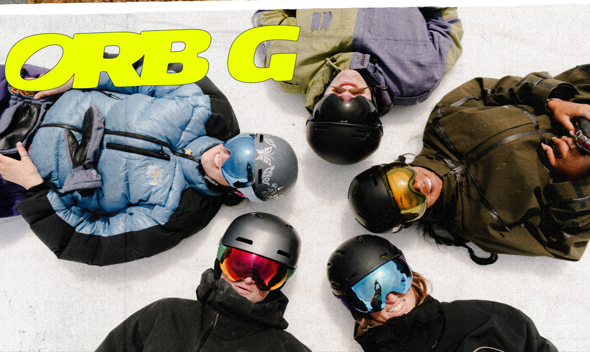 Five snowboarders lying in a circle on snow, viewed from above, with a large yellow 'ORB G' logo in the top left. They wear helmets, goggles, and colorful snowboard jackets in blue, olive green, black, and purple.