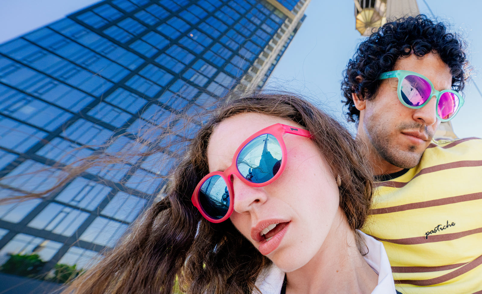 Two people wearing colorful sunglasses in an outdoor urban setting with a glass skyscraper in the background. The person in the foreground wears bright pink-framed sunglasses with mirrored blue lenses, and the person behind wears green-framed sunglasses with mirrored pink lenses.